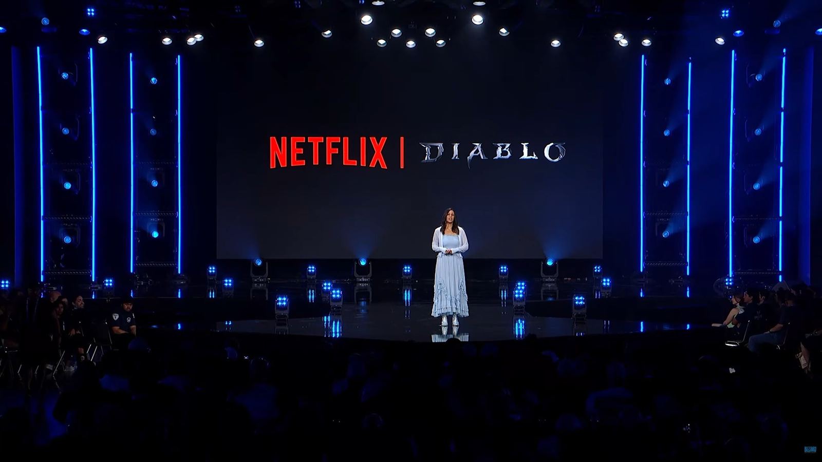 Escenario de la BlizzCon con una mujer de vestido claro hablando sola en el centro, ante una pantalla gigante que muestra el logotipo rojo de Netflix junto al logotipo de Diablo, con focos azules a los lados y público en penumbra