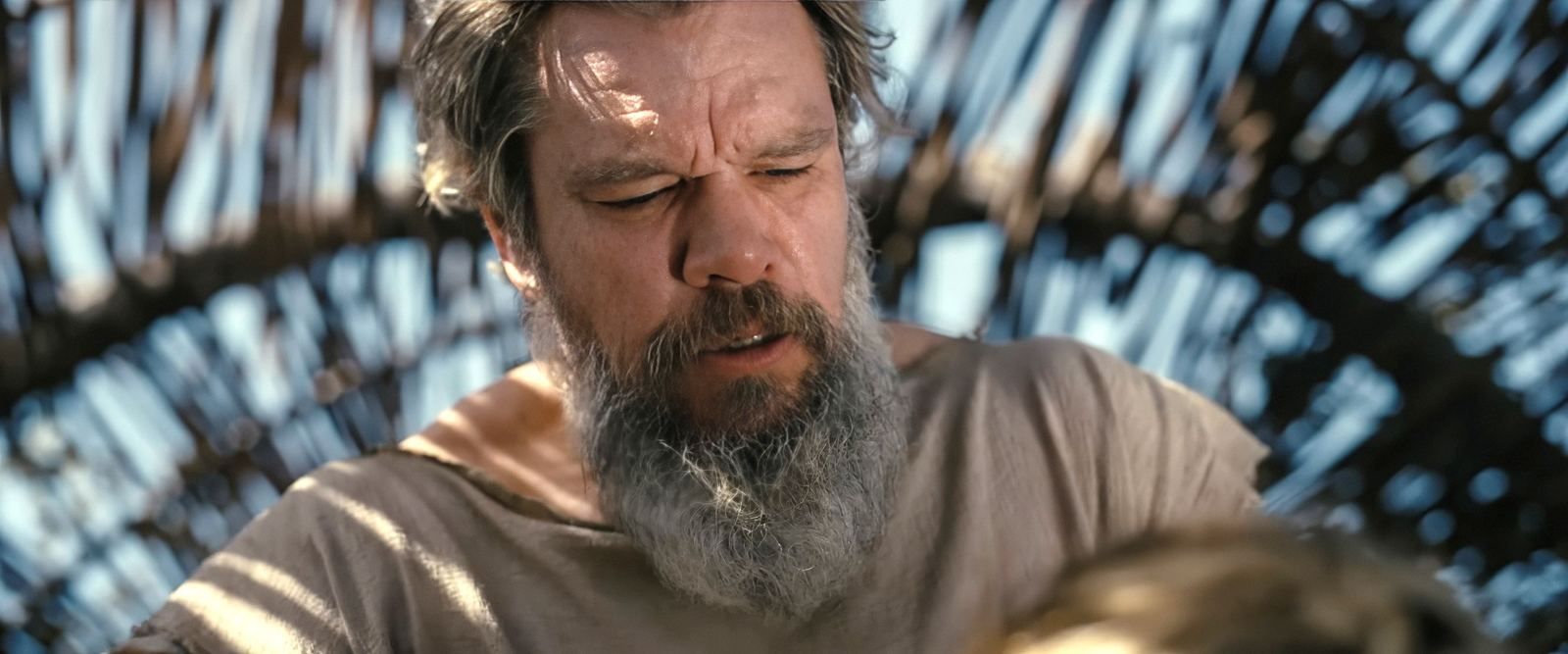 Matt Damon caracterizado como Odiseo, con barba canosa y túnica clara, entre sombras de luz filtrada por un entramado de madera
