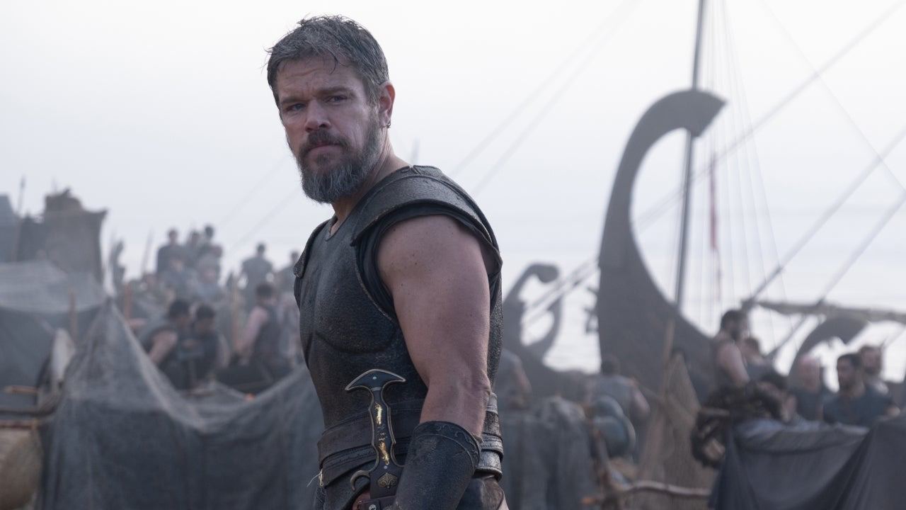 Matt Damon como Odiseo, con armadura de cuero y espada al cinto, de pie en una playa ante barcos griegos y soldados con redes