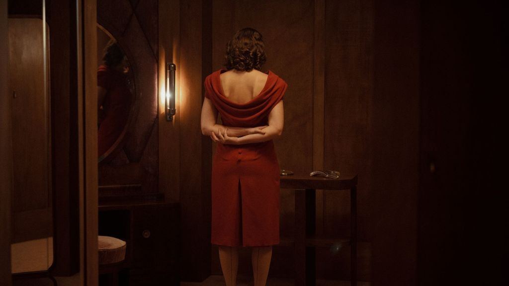 Fotograma de la película: una mujer de pelo corto ondulado, vestida de rojo, está de espaldas y con las manos juntas a la espalda en una habitación de paredes de madera en penumbra, junto a un espejo redondo y un aplique encendido