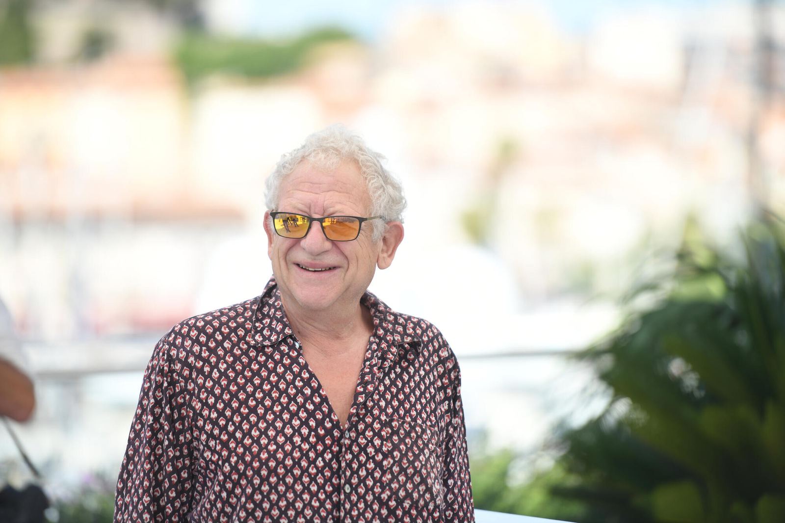 Jeremy Thomas, de pelo blanco y rizado y gafas de cristal ambarino, sonríe con camisa estampada en un pase de fotos al aire libre