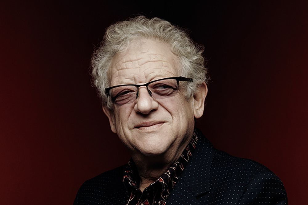 Retrato de estudio de Jeremy Thomas sobre fondo rojo oscuro, con gafas de montura negra y chaqueta azul marino sobre una camisa estampada